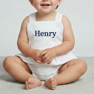 Baby wearing a white romper with 'Henry' printed on it, sitting on a light gray surface.